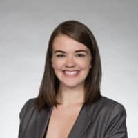 Photo of a woman with shoulder length straight brown hair and brown eyes. She is smiling. She is wearing a grey v neck blouse. The background is light grey.