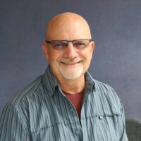 Photo of a man who is bald with light skin and a grey beard and mustache wearing glasses. He is wearing a collared striped blue shirt over a red t-shirt.