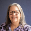 Photo of a woman with light skin and grey shoulder length straight hair wearing glasses. She is wearing a collared purple patterned shirt.