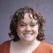 Photo of a fair skinned woman with blue eyes and brown curly shoulder length hair wearing a burnt orange asymmetrical shirt.