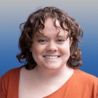 Photo of a woman with brown curly shoulder length hair and blue eyes smiling. She is wearing a dark orange t-shirt with an asymmetrical neckline.