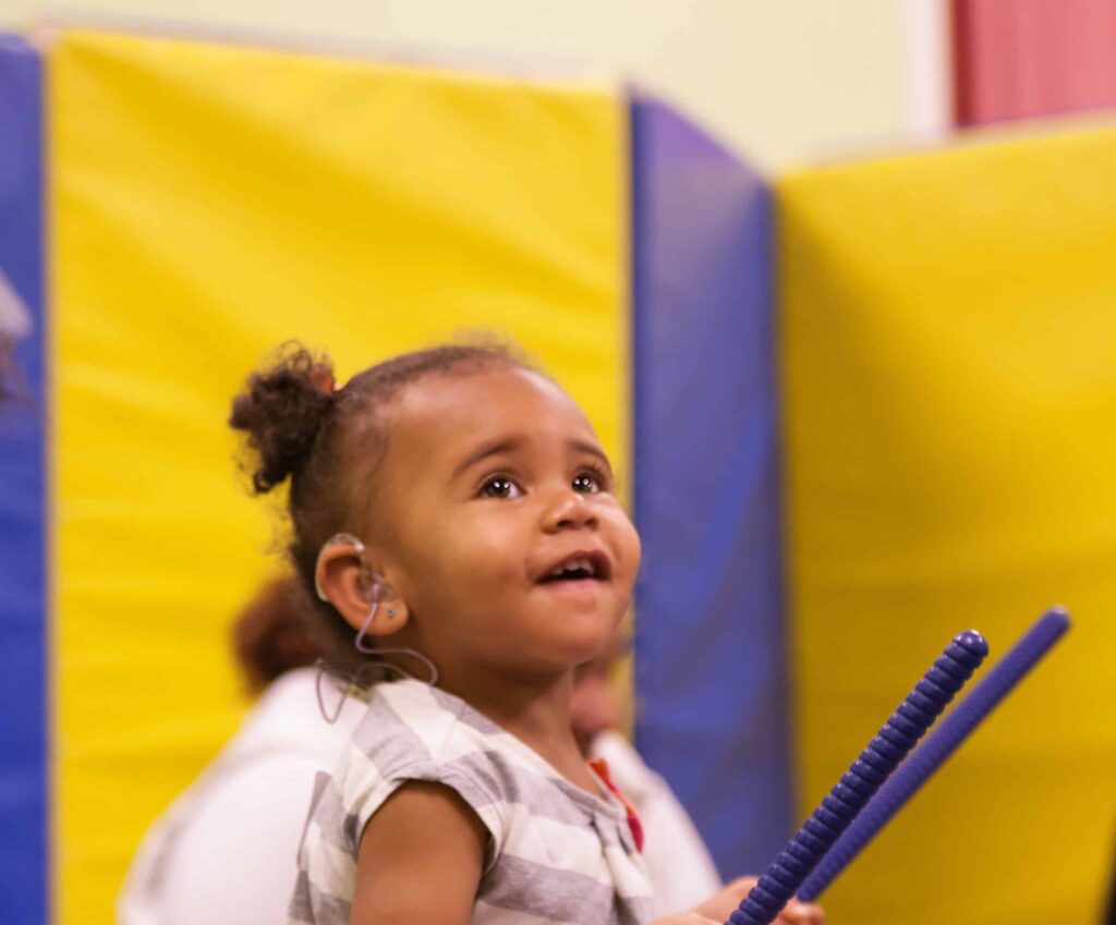 Child smiles while playing sticks and listening to music at Music and Movement at Listen and Talk.