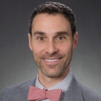 Photo of a man with short dark hair and blue eyes smiling. He is wearing a red checked bow tie with a blue and white checked collared shirt and grey suit jacket.