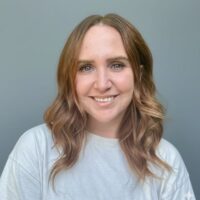 Photo of a woman with long wavy light auburn hair with light skin and blue eyes. She is smiling. She is wearing a white long sleeved t-shirt.