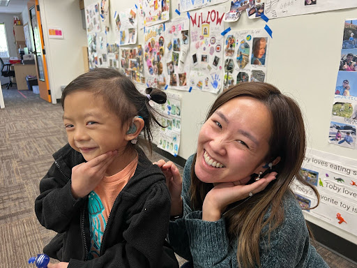 Listen and Talk's Audiologist Christie poses with a child who proudly shows her hearing aid in front of the camera!