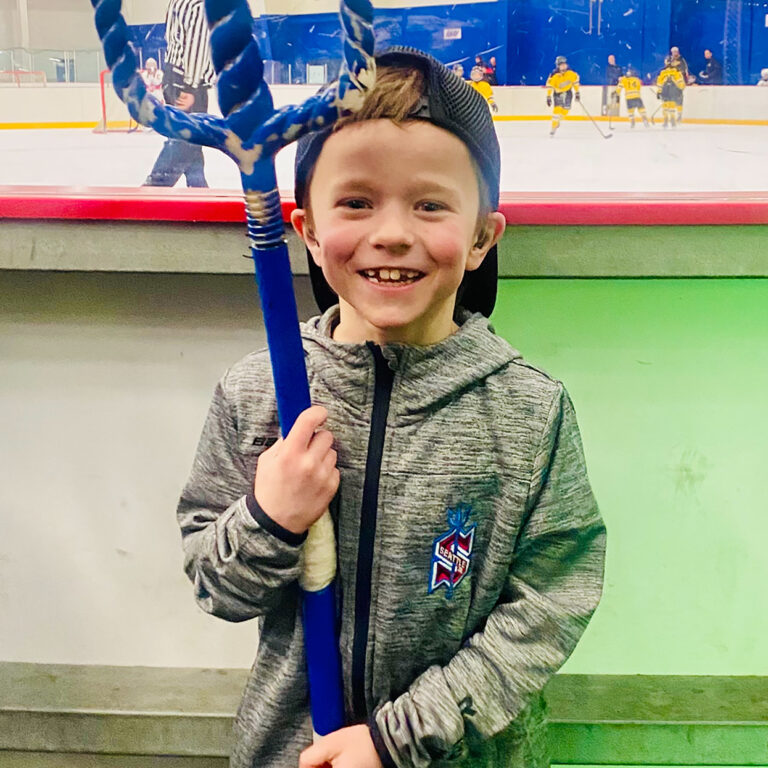 Alum Caleb stands in front of a hockey rink smiling for the camera as he holds a blue trident stick. He has a blue baseball cap on backwards and wears a long-sleeve grey zipped up hoodie on with the Seattle Kraken logo.