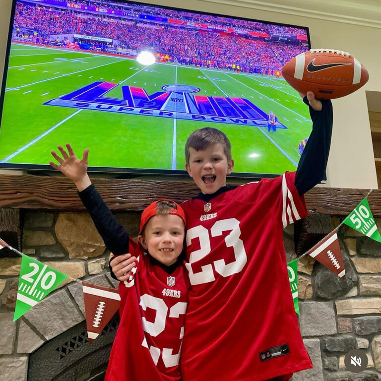 Caleb and his older brother smile for the camera wearing red football 49ers jerseys as they stand in front of a large TV screen showing the latest Superbowl Game. Both of them have their hands up in the air while his brother holds a football.