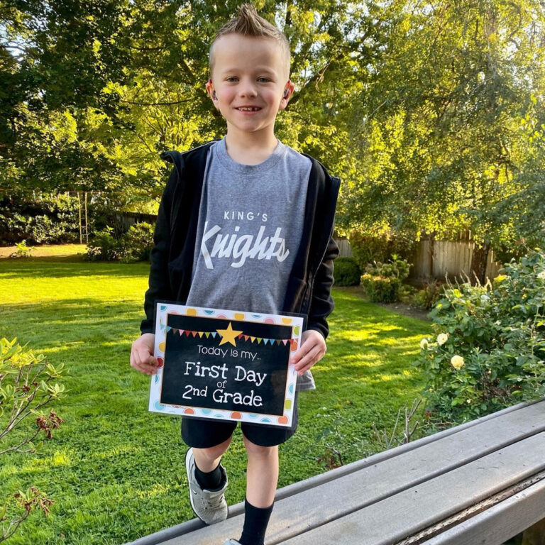 Caleb holds a "today is my first day of second grade sign" while standing on a wooden bench in front of a park with grass and trees.