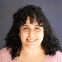 Photo of a woman with fair skin and black curly shoulder length hair wearing a pink blouse.