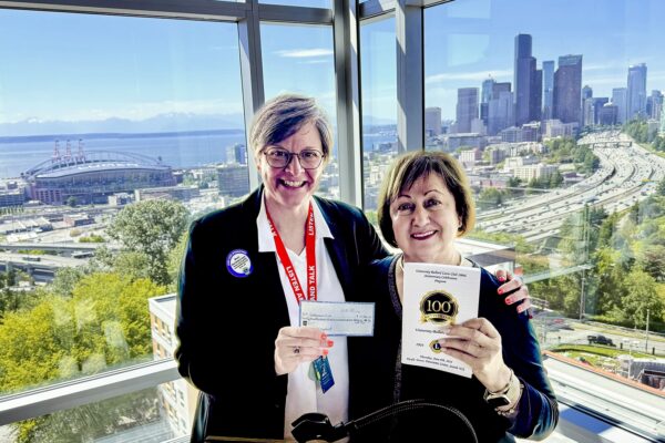 Executive Director Maura Berndsen stands next to University-Ballard Lions Club President, Norah Giraldo as they stand in front of a large panoramic window featuring a scenic view of iconic Seattle landmarks including Alki Beach in West Seattle, downtown Seattle, the Olympic Mountains, Lumen Field, and I-5. Berndsen holds up a check while Giraldo holds up a program book.