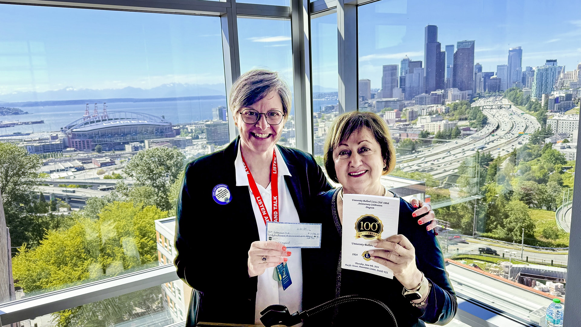 Executive Director Maura Berndsen stands next to University-Ballard Lions Club President, Norah Giraldo as they stand in front of a large panoramic window featuring a scenic view of iconic Seattle landmarks including Alki Beach in West Seattle, downtown Seattle, the Olympic Mountains, Lumen Field, and I-5. Berndsen holds up a check while Giraldo holds up a program book.