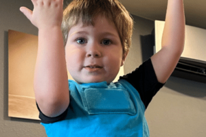 Young boy with hands up above his head wearing a light blue LENA vest