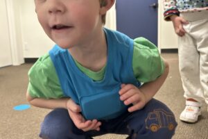 Young boy with light brown hair and blue eyes wearing a blue LENA vest over a green t-shirt