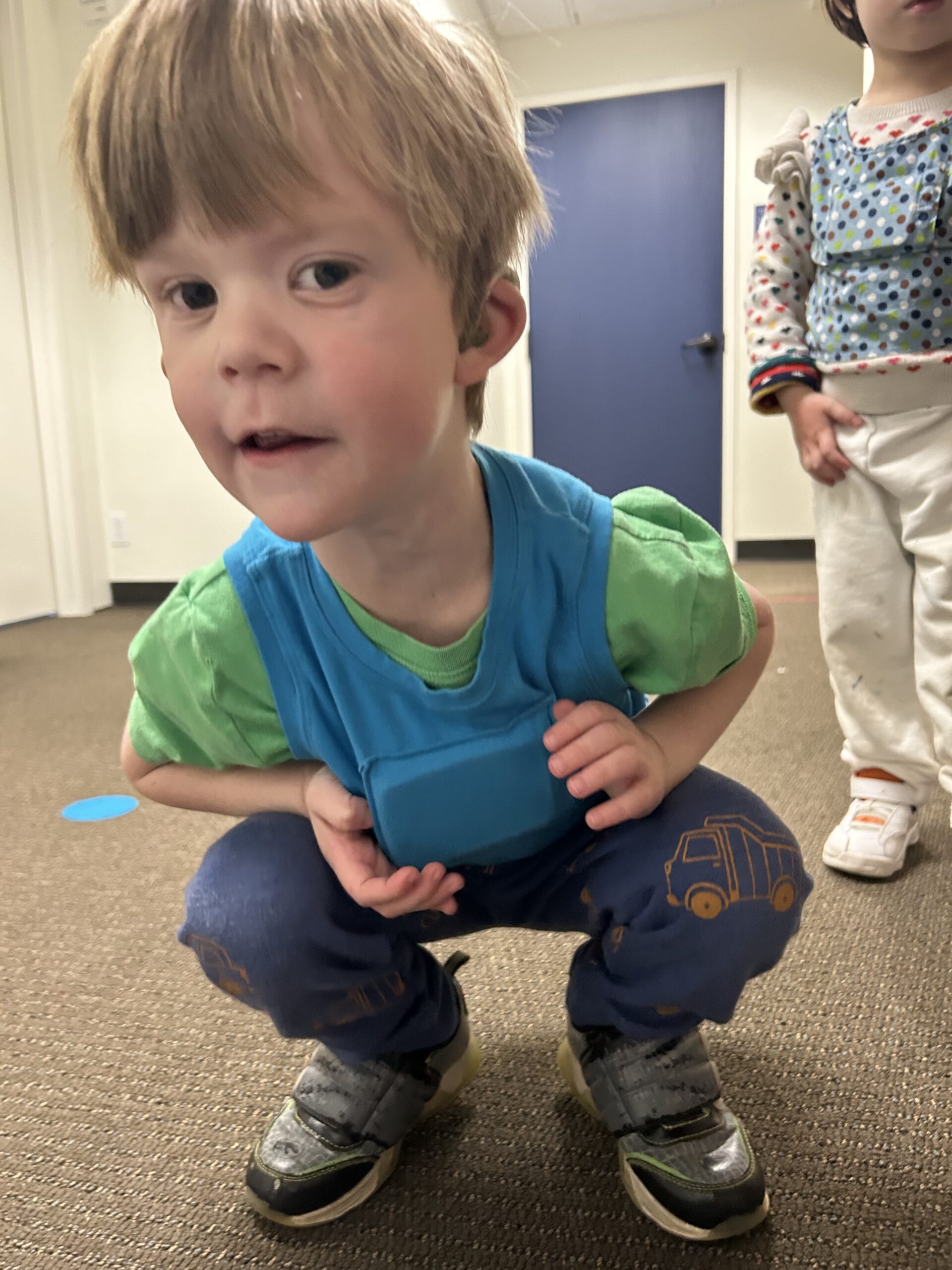 Young boy with light brown hair and blue eyes wearing a blue LENA vest over a green t-shirt