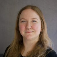 Photo of a woman with blue eyes and long straight dark blonde hair. She has a nose ring and is smiling.