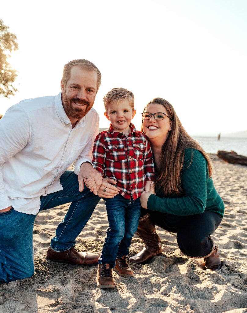 Russell Family Photo 1 Man with light brown hair and beard and mustache wearing a white button up shirt kneels on his right knee. A preschool aged boy with blonde hair and blue eyes is wearing a button up plaid blue, red and white shirt is standing to the right of the man. A woman with long straight brown hair wearing a teal colored sweater. The people are standing on sand with water visible in the background on the right.