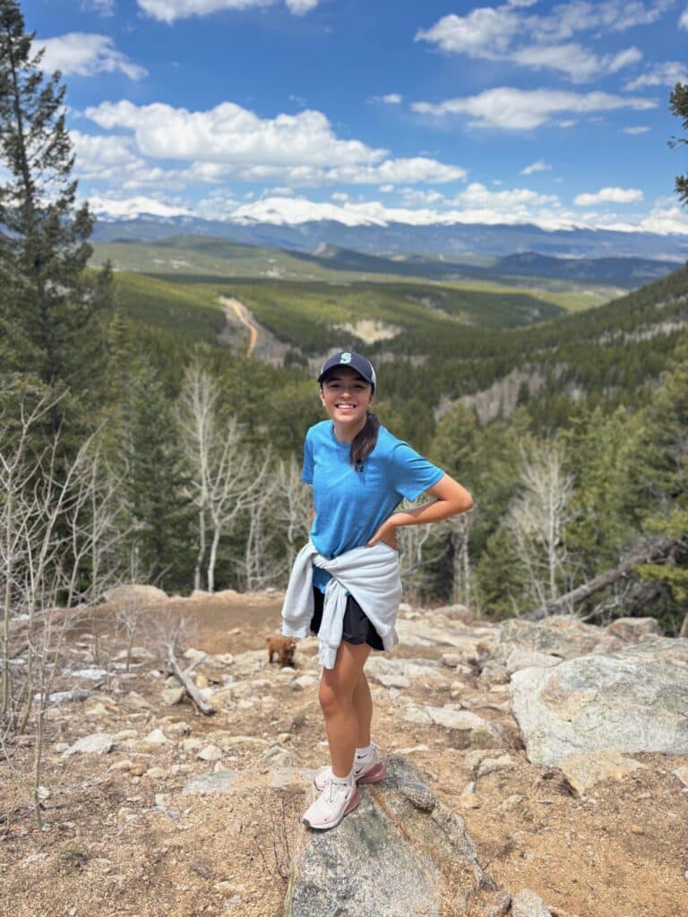 Avy teenager hiking Photo of a teen girl wearing a light blue t-shirt and black shorts with a navy baseball cap on her head. She has a brown ponytail and brown eyes. In the background is a blue sky with white clouds and a treeline. She is standing on a rock in beige dirt.