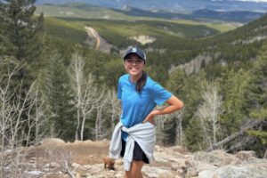 Photo of a teen girl wearing a light blue t-shirt and black shorts with a navy baseball cap on her head. She has a brown ponytail and brown eyes. In the background is a blue sky with white clouds and a treeline. She is standing on a rock in beige dirt.