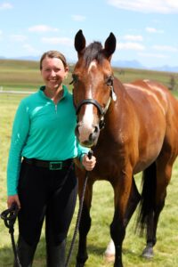 Photo of a woman with brown hair pulled back from her face on the left side of the photo. She is wearing a light green long sleeved shirt and black riding pants holding the rein of a horse that stands to her right. The horse is brown with a white streak on its nose. Light blue sky is above the green grass they stand on.