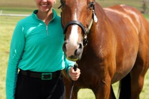 Photo of a woman with brown hair pulled back from her face on the left side of the photo. She is wearing a light green long sleeved shirt and black riding pants holding the rein of a horse that stands to her right. The horse is brown with a white streak on its nose. Light blue sky is above the green grass they stand on.