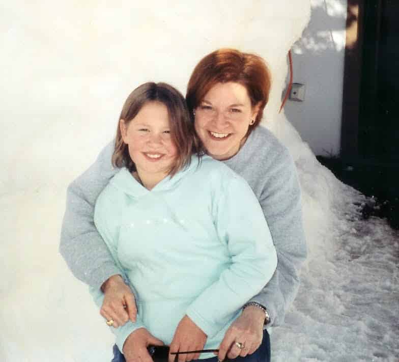 Photo of a school aged girl with brown straight hair wearing a light teal sweatshirt. To her right is a woman with short auburn hair wearing a gray sweater with her arms surrounding the girl There is a white snowbank in the background.