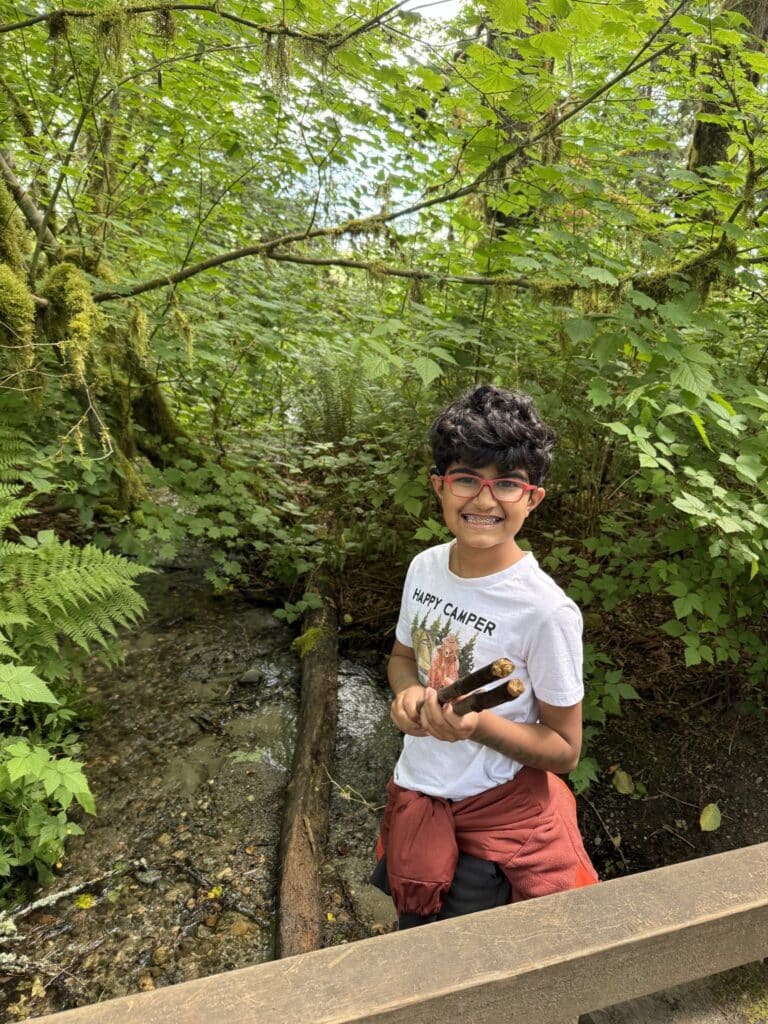 y4O6YT0oTCmKbDp1_DTh6Q Photo of an eight year old boy with dark wavy hair and red framed glasses. He is wearing a white t-shirt with red shorts and holding two sticks. There is a green leafy tree in a forest in the background.