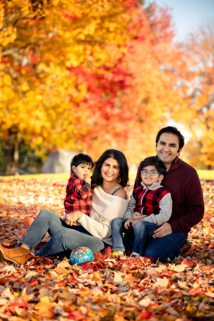 zane4 Photo of a family in a fall scene sitting on a bed of leaves that are orange, yellow and red. A young boy with dark brown hair wearing a red and black plaid shirt is on the left in his mother's arms. The woman has shoulder length straight dark brown hair and wears a off the shoulder cream colored sweater. To their right is a boy who is a bit older with dark brown hair and glasses wearing a red and black plaid vest over a grey shirt sitting on his father's lap. HIs father has dark brown short hair and wears a burgundy sweater. All family members are smiling.