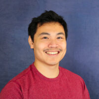 Photo of a man with short black hair and mustache smiling. He is wearing a red t-shirt.