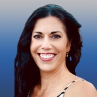 Photo of a woman with short blonde hair and blue eyes. She is wearing white horn rimmed glasses and smiling. She wears a white lace blouse.