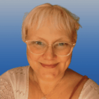 Photo of a woman with short straight blonde hair and bue eyes. She is wearing light colored cat-eye shaped glasses andd is smiling. She wears white lacy blouse.