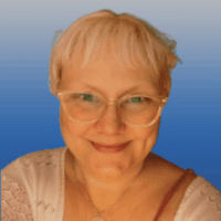 Photo of a woman with short blonde hair and blue eyes. She is wearing white horn rimmed glasses and smiling. She wears a white lace blouse.