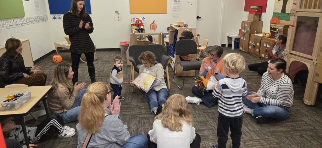 A woman with curly brown hair sits on the floor in the center of the room reading a book out loud to a child who is walking up to her. Other adults and children sit in a circle looking on.