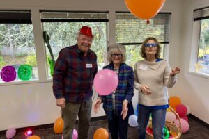 Photo of a man wearing a red hat on the left, a woman with grey hair wearing black framed glasses and a silver hat in the middle and a woman with curly brown hair and star shaped blue glasses on the right. Orange balloons are on the floor. There is one big orange balloon above the woman with curly hair and a pink balloon in between the man and the woman,