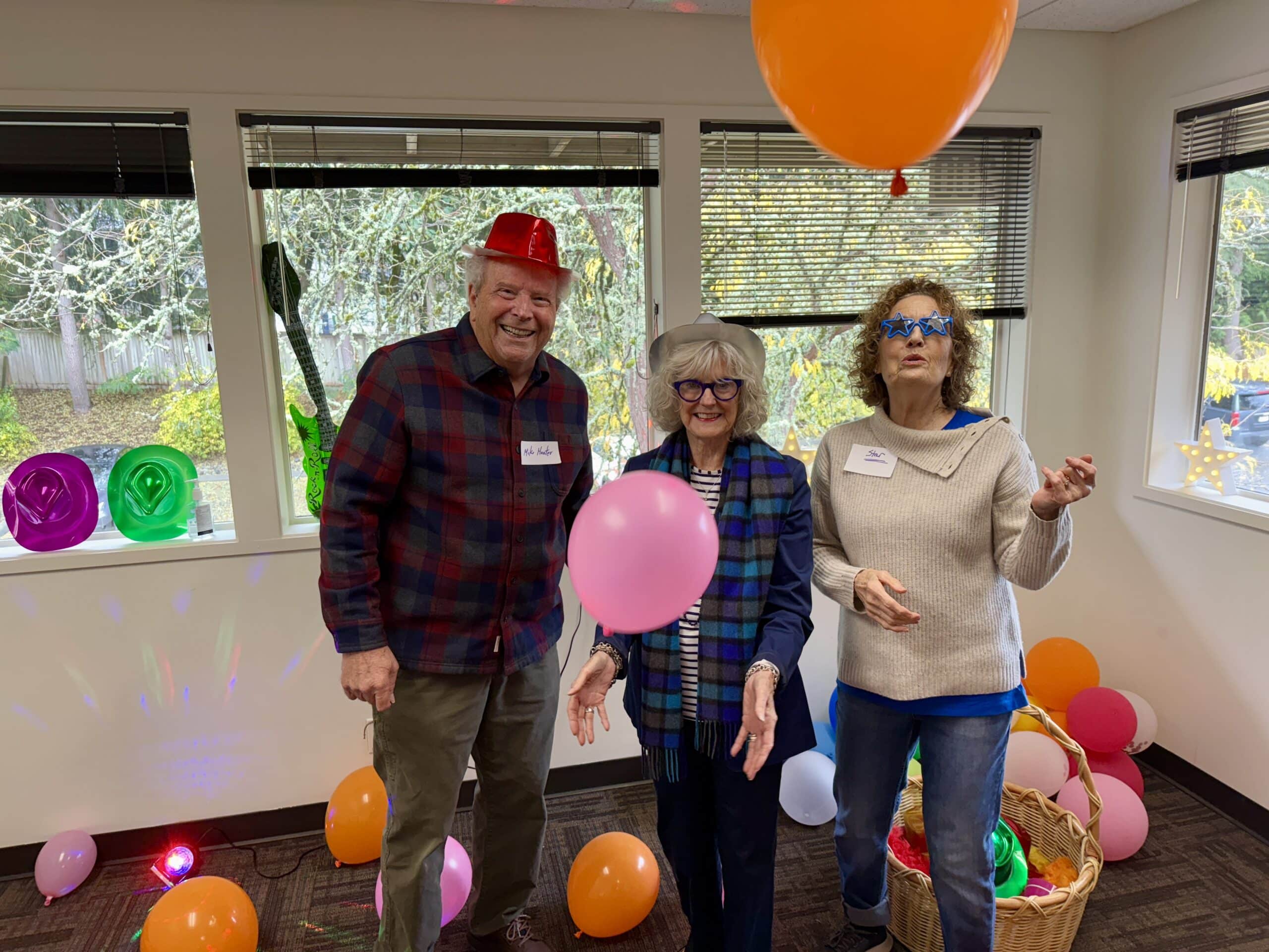 Photo of a man wearing a red hat on the left, a woman with grey hair wearing black framed glasses and a silver hat in the middle and a woman with curly brown hair and star shaped blue glasses on the right. Orange balloons are on the floor. There is one big orange balloon above the woman with curly hair and a pink balloon in between the man and the woman,