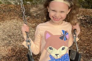 Photo of a girl on a swing smiling. She is wearing a t-shirt with a fox on it. and striped pants with blue sneakers. She wears a headband.