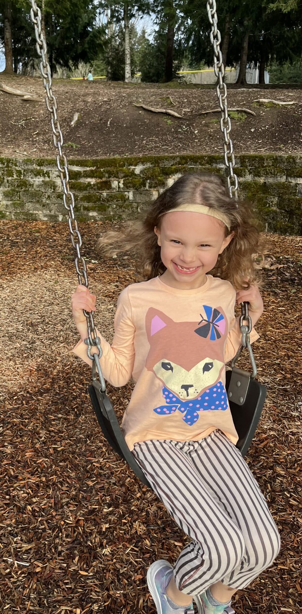 Photo of a girl on a swing smiling. She is wearing a t-shirt with a fox on it. and striped pants with blue sneakers. She wears a headband.