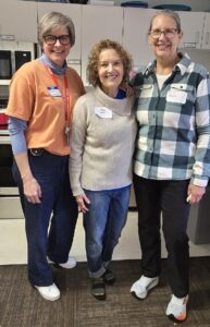 Photo of three women smiling. The woman on the left is wearing an orange t-shirt and blue jeans and has short grey hair and black-framed glasses. The woman in the middle is wearing a grey cowl necked sweater and blue jeans and has short curly brown hair. The woman on the right is tall and has grey hair pulled back. She is wearing a green and white checked button up shirt and black pants.