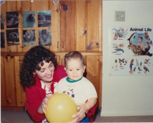 A woman with curly brown hair wearing a read cardigan sweater smiles while holding a yellow balloon with a preschool aged boy. The boy is wearing a white t-shirt with a green crewneck. There is wood paneling in the background with children's art on the walls.