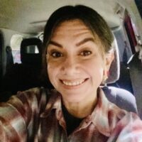 Photo of a woman with dark hair pulled back from her face smiling. She is wearing a red and gray checked collared shirt. She is shown in a car.