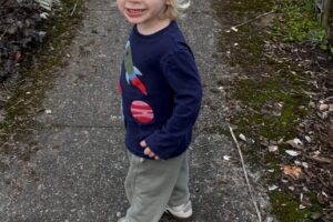 Photo of a preschool aged boy with shoulder length blonde hair. He is wearing a light blue knit hat with a pom pom on top and a navy blue sweater with grey pants and white sneakers. He is standing outside on a sidewalk with plants lining the path.