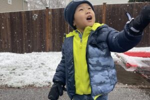 Photo of a preschool aged boy wearing a navy blue hat and puffy jacket with a light green fleece pullover. He is wearing jeans and snow boots. There is snow in the background and he is holding his mittened hand up to the sky while he looks up.