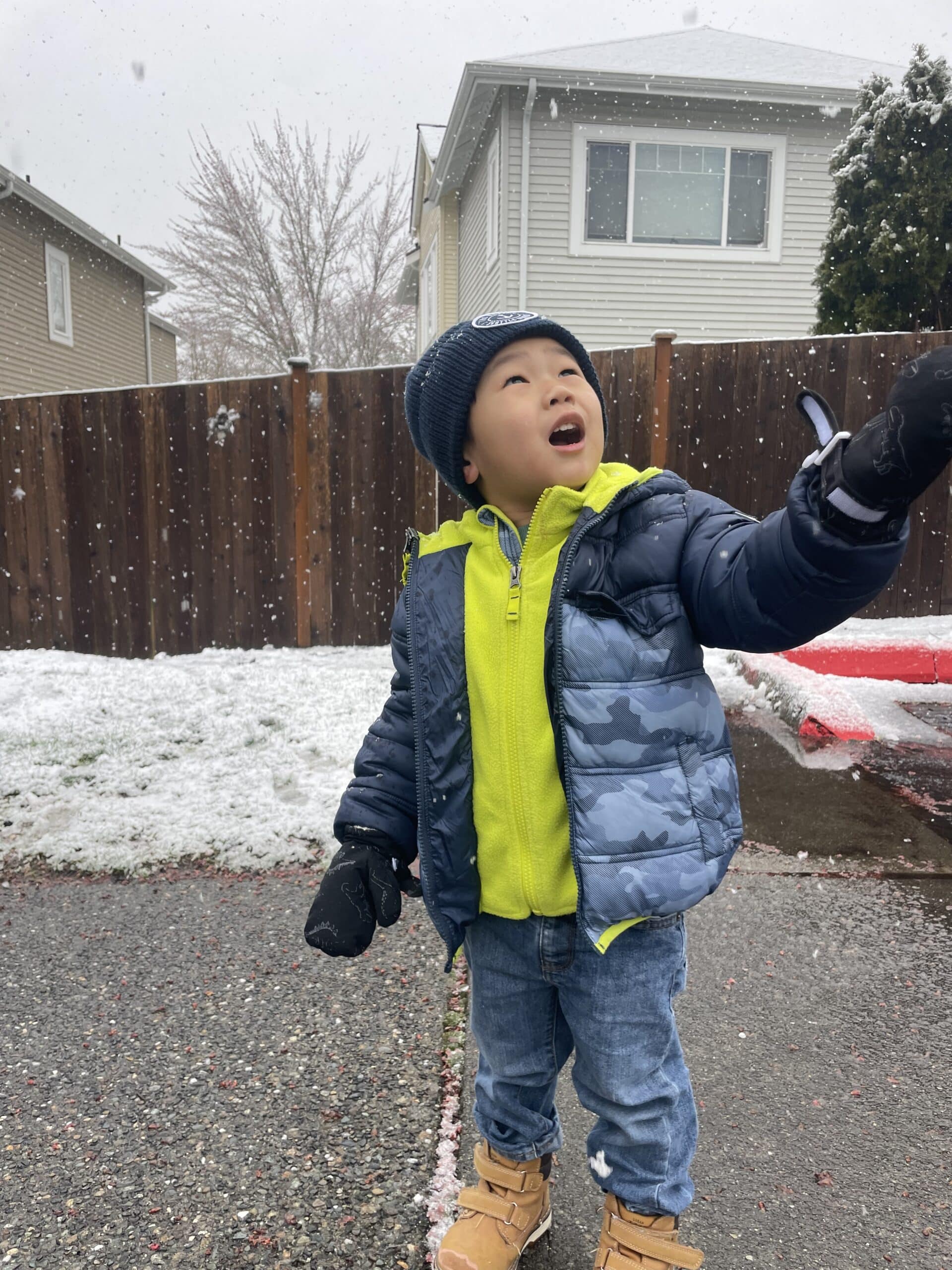 Photo of a preschool aged boy wearing a navy blue hat and puffy jacket with a light green fleece pullover. He is wearing jeans and snow boots. There is snow in the background and he is holding his mittened hand up to the sky while he looks up.
