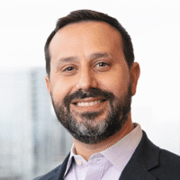Photo of a man with dark short hair and a beard and mustache smiling. He is wearing a lilac colored collared shirt and a dark suit jacket.