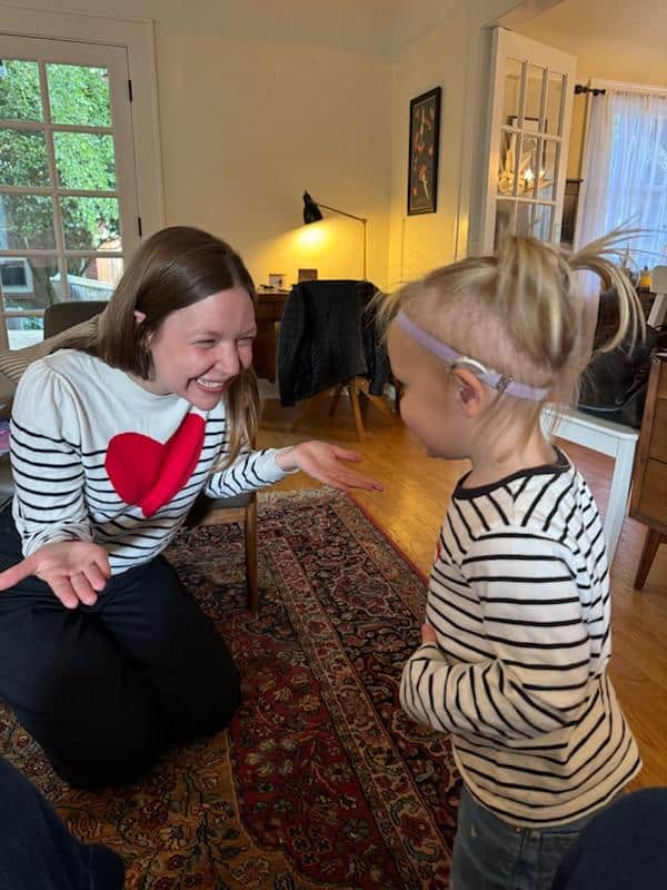 FrMcB 1 Photo of a child on the right wearing hearing technology on her left ear. She has blonde pigtails and is looking at a woman with long brown hair on the left who is smiling and wearing a sweater with a big red heart on it.