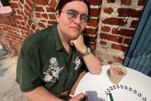 Photo of a young man wearing glasses with his hair pulled back from his face. His left elbow rests on a white table. He is holding a black pen in his right hand poised on a notebook. He is wearing a button up green short sleeved shirt with a beige design and brown pants. There is a brick wall in the background.