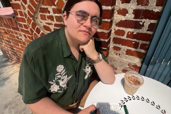 Photo of a young man wearing glasses with his hair pulled back from his face. His left elbow rests on a white table. He is holding a black pen in his right hand poised on a notebook. He is wearing a button up green short sleeved shirt with a beige design and brown pants. There is a brick wall in the background.