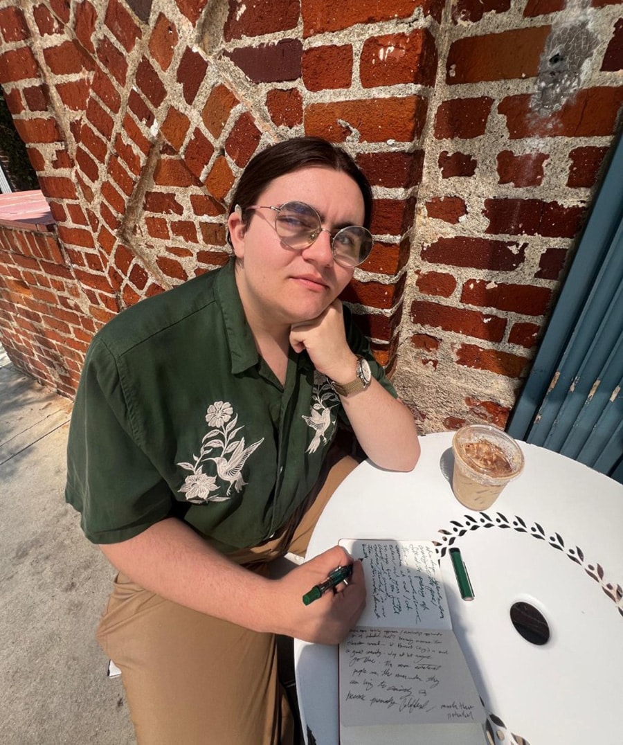 Photo of a young man wearing glasses with his hair pulled back from his face. His left elbow rests on a white table. He is holding a black pen in his right hand poised on a notebook. He is wearing a button up green short sleeved shirt with a beige design and brown pants. There is a brick wall in the background.