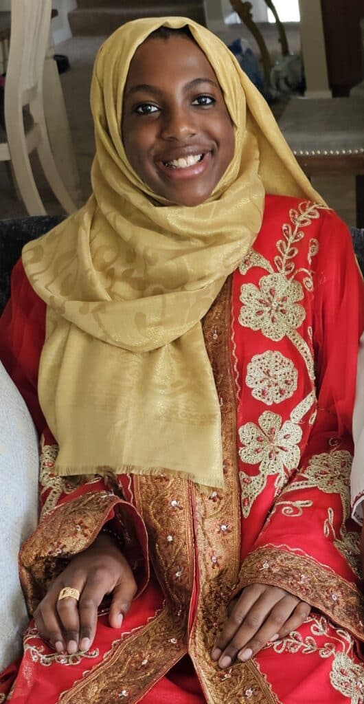 Photo of a woman with brown skin, brown eyes and black hair smiling wearing a gold fabric hijab and red patterned dress.