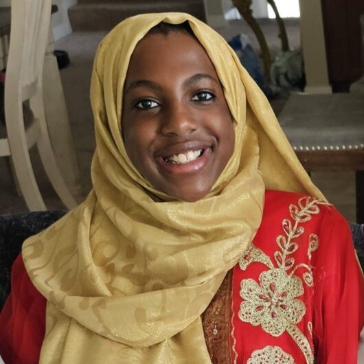 Amalia3 (1) Photo of a woman with brown skin, brown eyes and black hair smiling wearing a gold fabric hijab and red patterned dress.