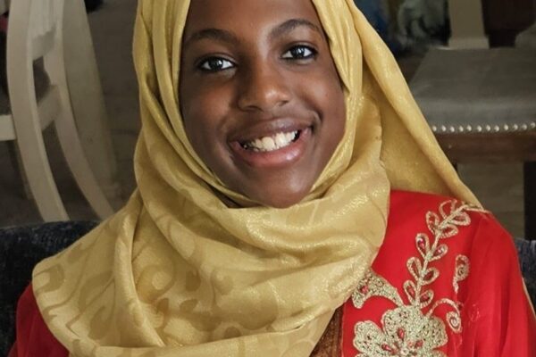 Photo of a woman with brown skin, brown eyes and black hair smiling wearing a gold fabric hijab and red patterned dress.
